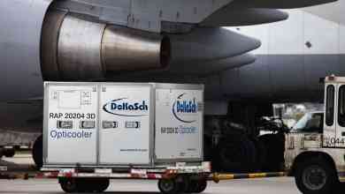 A large cooler on wheels for medicine transport sits on tarmac beneath an airplane wing. Air transport is commonly used to ship medicines.