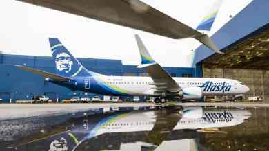 A white 737 MAX with Native Alaskan logo on tail sits in front of a hanger on wet pavement. Ground view from the side.