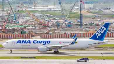 A white All Nippon Airways cargo plane with blue tail moves down a taxiway at Bangkok airport, with construction in the background.