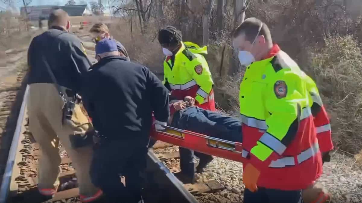 A photograph of police and ambulance workers lifting a man on a stretcher. They are next to train tracks.