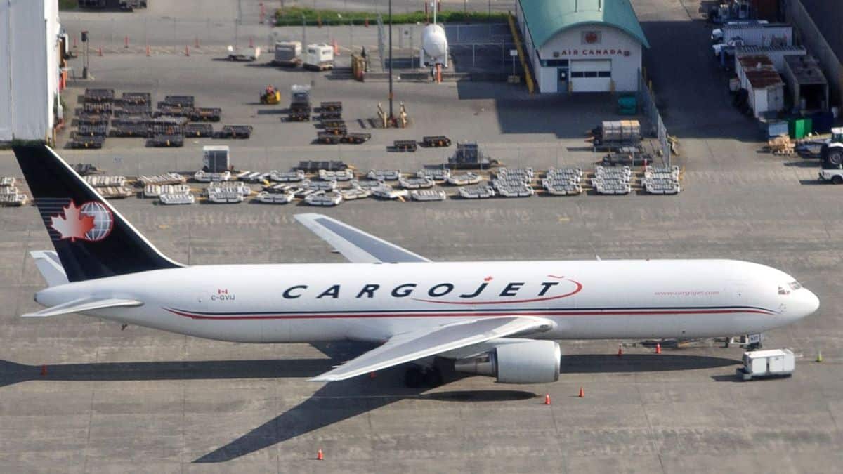 White cargo jet with blue tail sitting in front of cargo hangar. View from above in plane taking off.