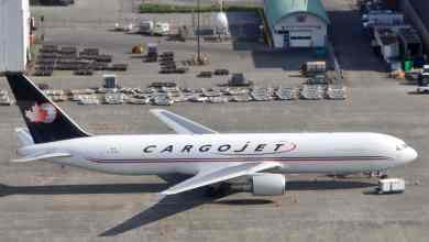 White cargo jet with blue tail sitting in front of cargo hangar. View from above in plane taking off.
