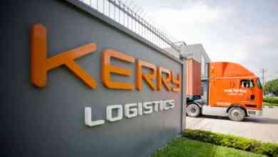 An orange Kerry Logistics truck entering road from Kerry facility next to company sign.