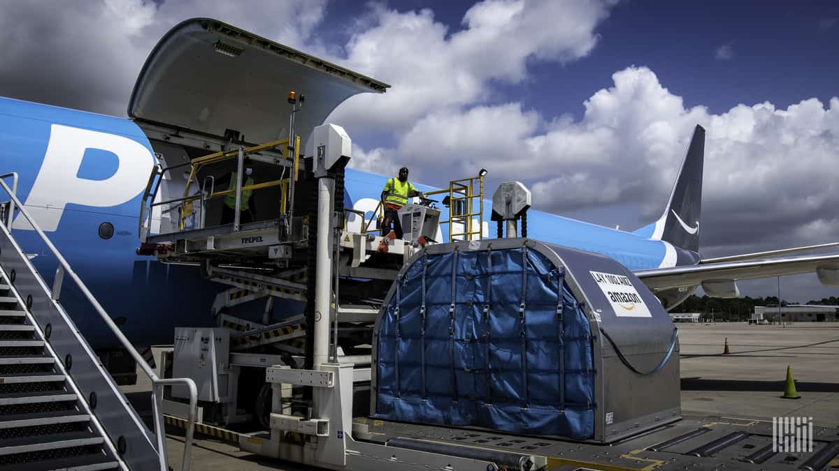 A light blue Amazon plane with side cargo door open and large metal container on the ground ready to be lifted for loading.