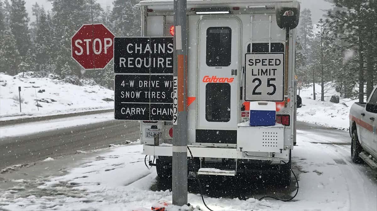 Highway department truck with "Chains Required" sign along a snowy California road.