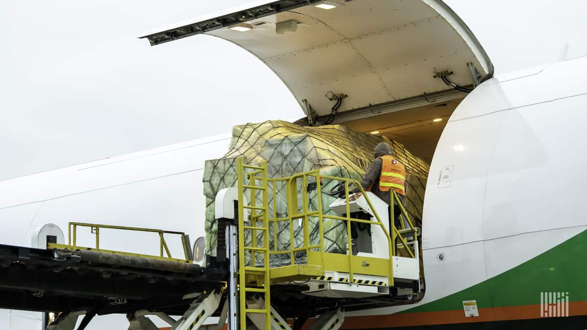 Cargo pallets on a hydraulic lift being unloaded out of side door of large aircraft with white fuselage.