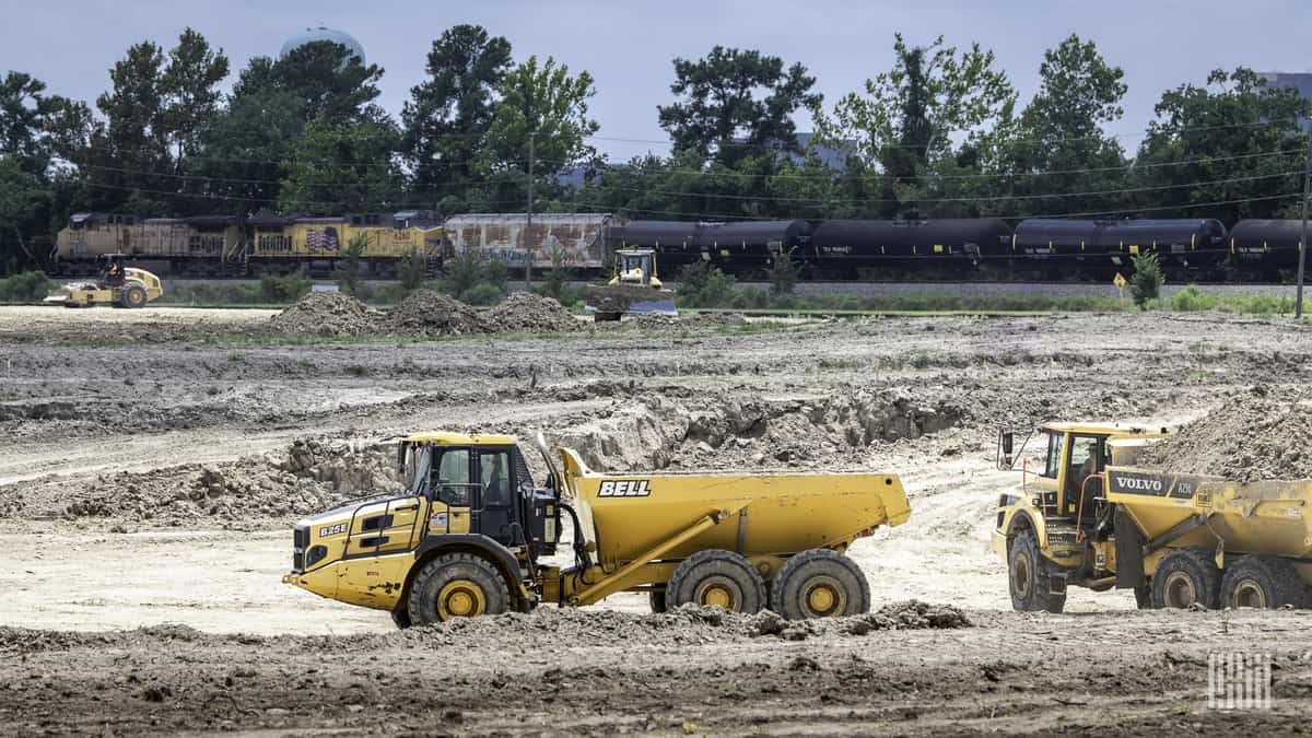 A photograph of a freight train passing through a field while a construction vehicle is in the field.