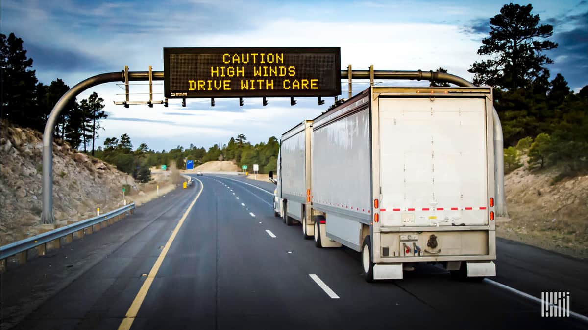 Tractor-trailer with "Caution: High Winds" highway sign above it.