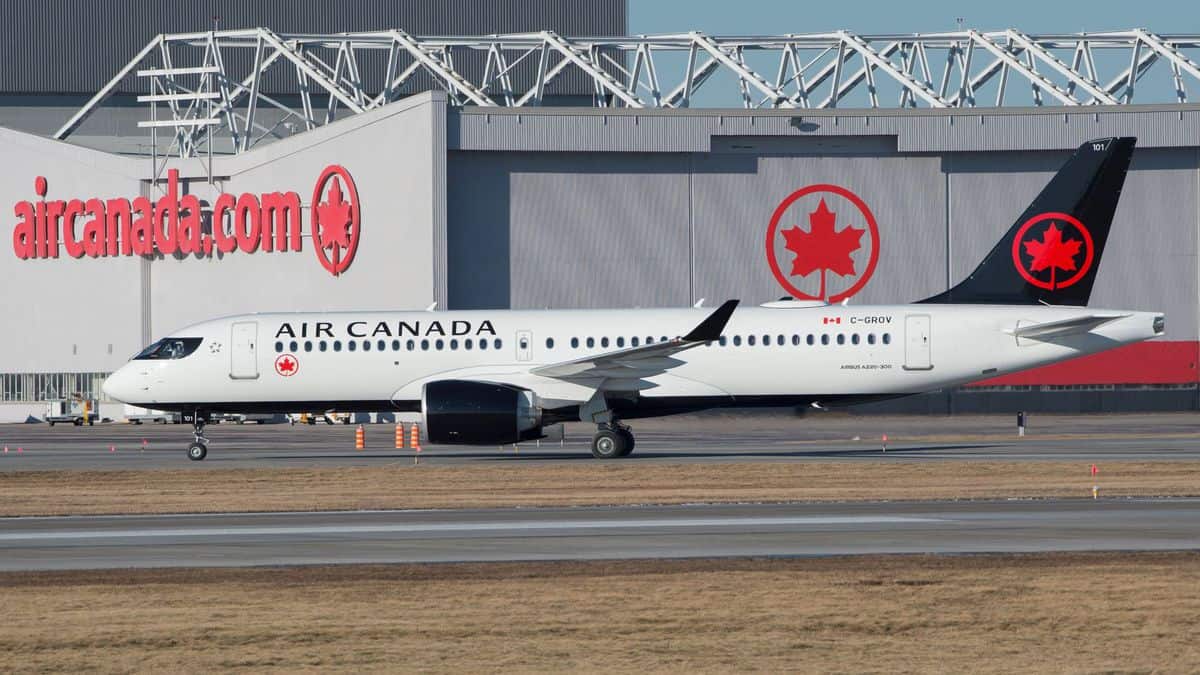 A white Air Canada jet with blue tail taxis in front of large aircraft hangar.