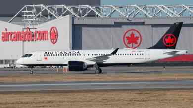 A white Air Canada jet with blue tail taxis in front of large aircraft hangar.
