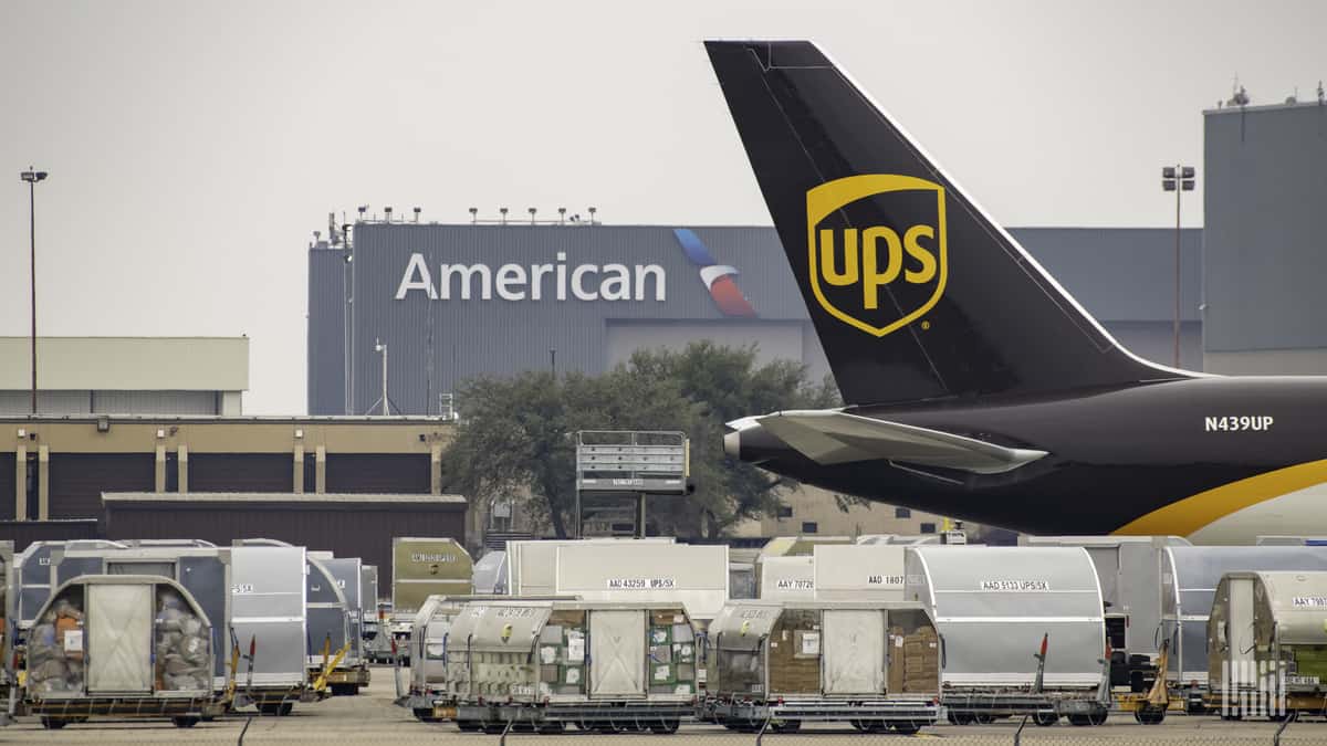 Brown tail of UPS jet in foreground, cargo containers and American Airlines cargo warehouse in background.