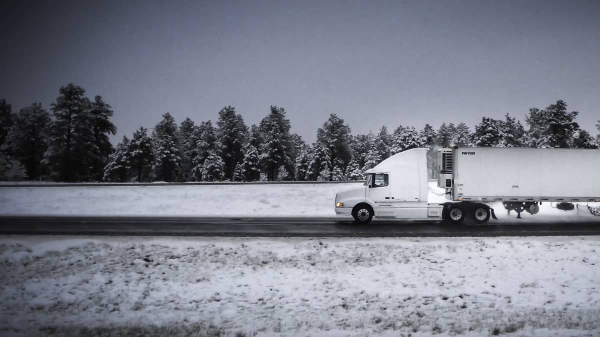 Tractor-trailer heading down highway with some snow on the road and surrounding hillsides.