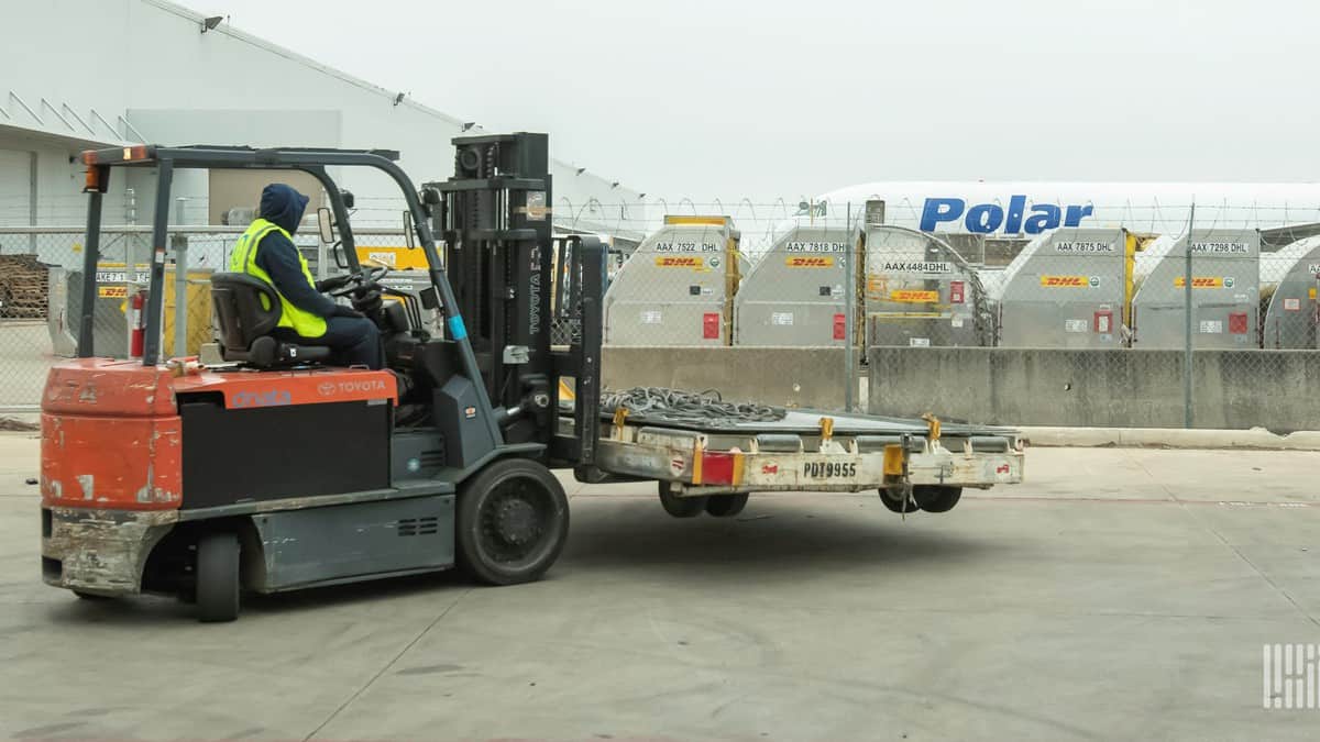 A forklift driver with a pallet, airfreight containers in a row and an aircraft in the background.