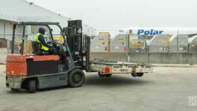A forklift driver with a pallet, airfreight containers in a row and an aircraft in the background.