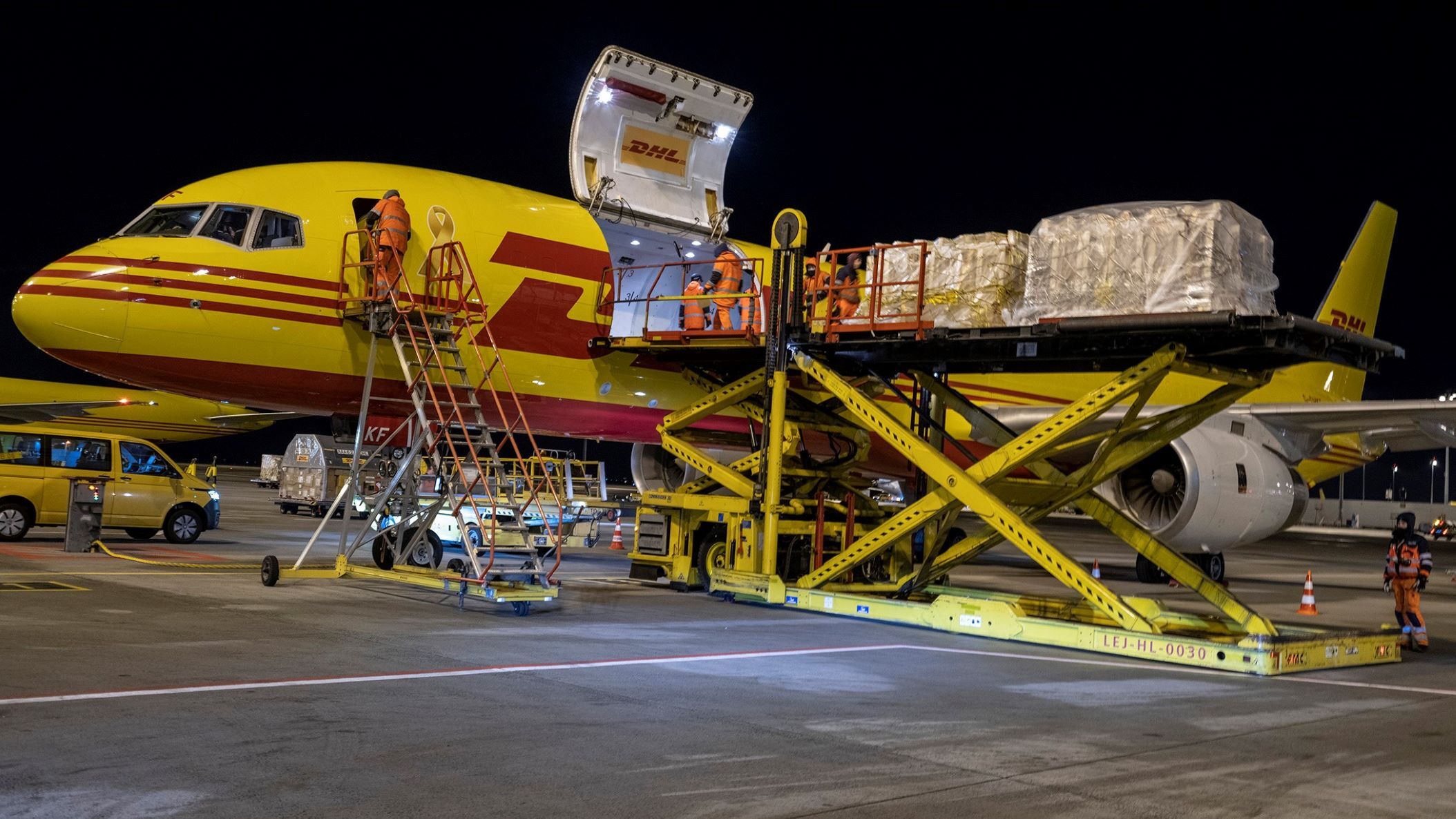 A mustard DHL cargo jet with red lettering loaded with cargo at night.