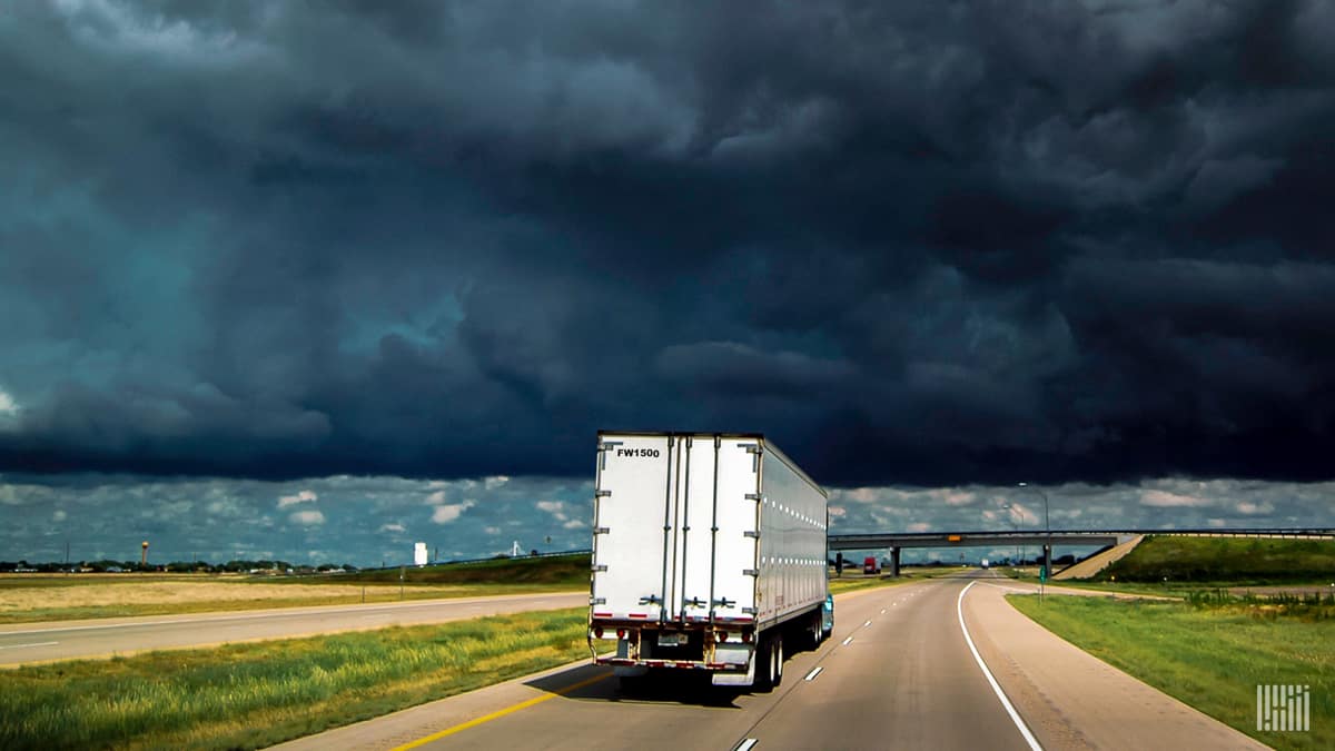 Tractor-trailer heading down a highway with dark thunderstorm cloud across the sky.