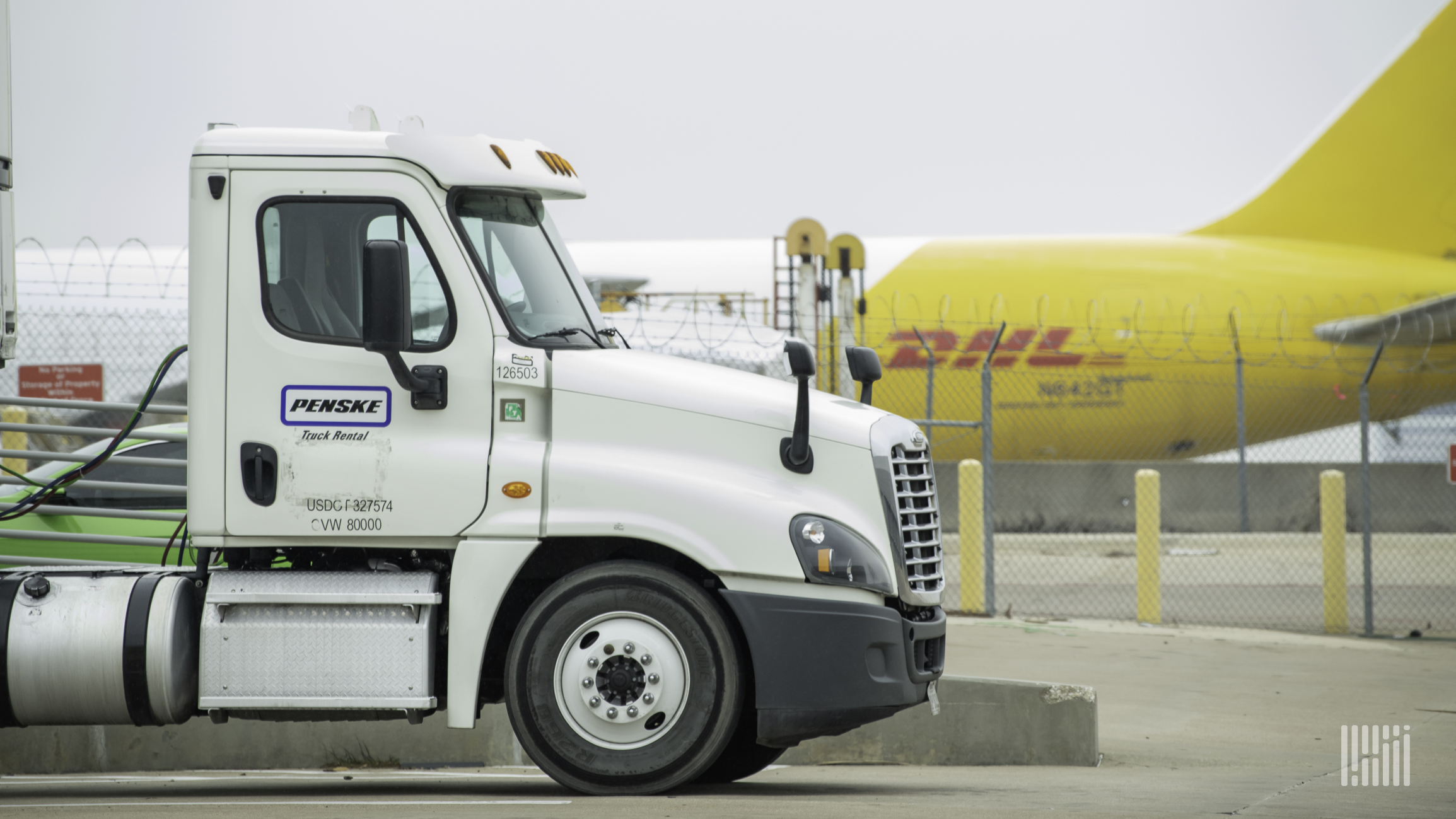 Back half of a mustard-colored DHL plane in background and tractor-trailer cab in foreground at airport.