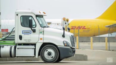 Back half of a mustard-colored DHL plane in background and tractor-trailer cab in foreground at airport.