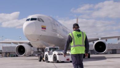 An airport worker with a yellow vest walks towards an airplane and a ground tug.