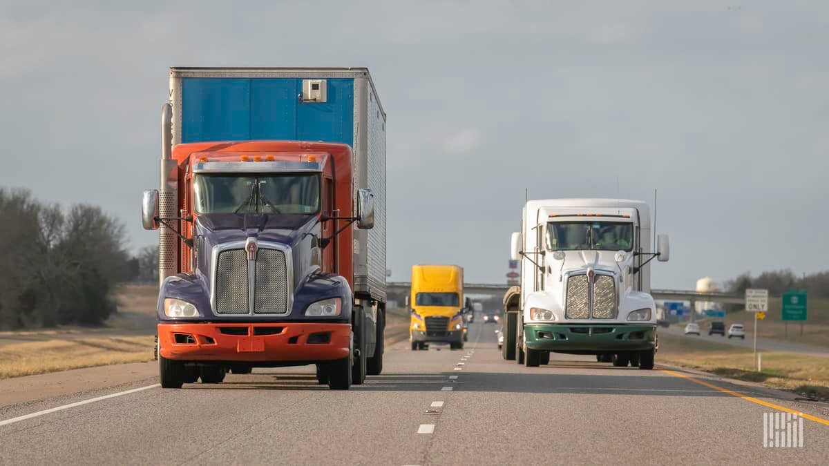 Three semi-trucks travel on a highway, seen from the front at varying distances.