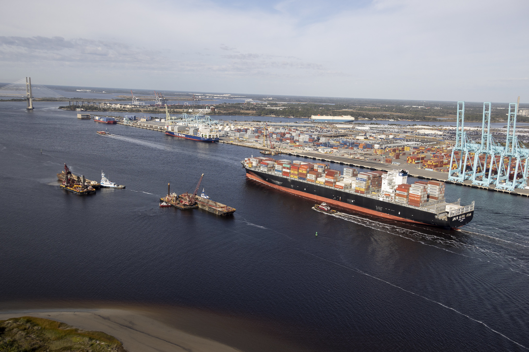 A photograph of a container vessel passing through a shipping channel. There are two smaller boats next to the vessel.