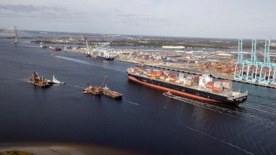 A photograph of a container vessel passing through a shipping channel. There are two smaller boats next to the vessel.
