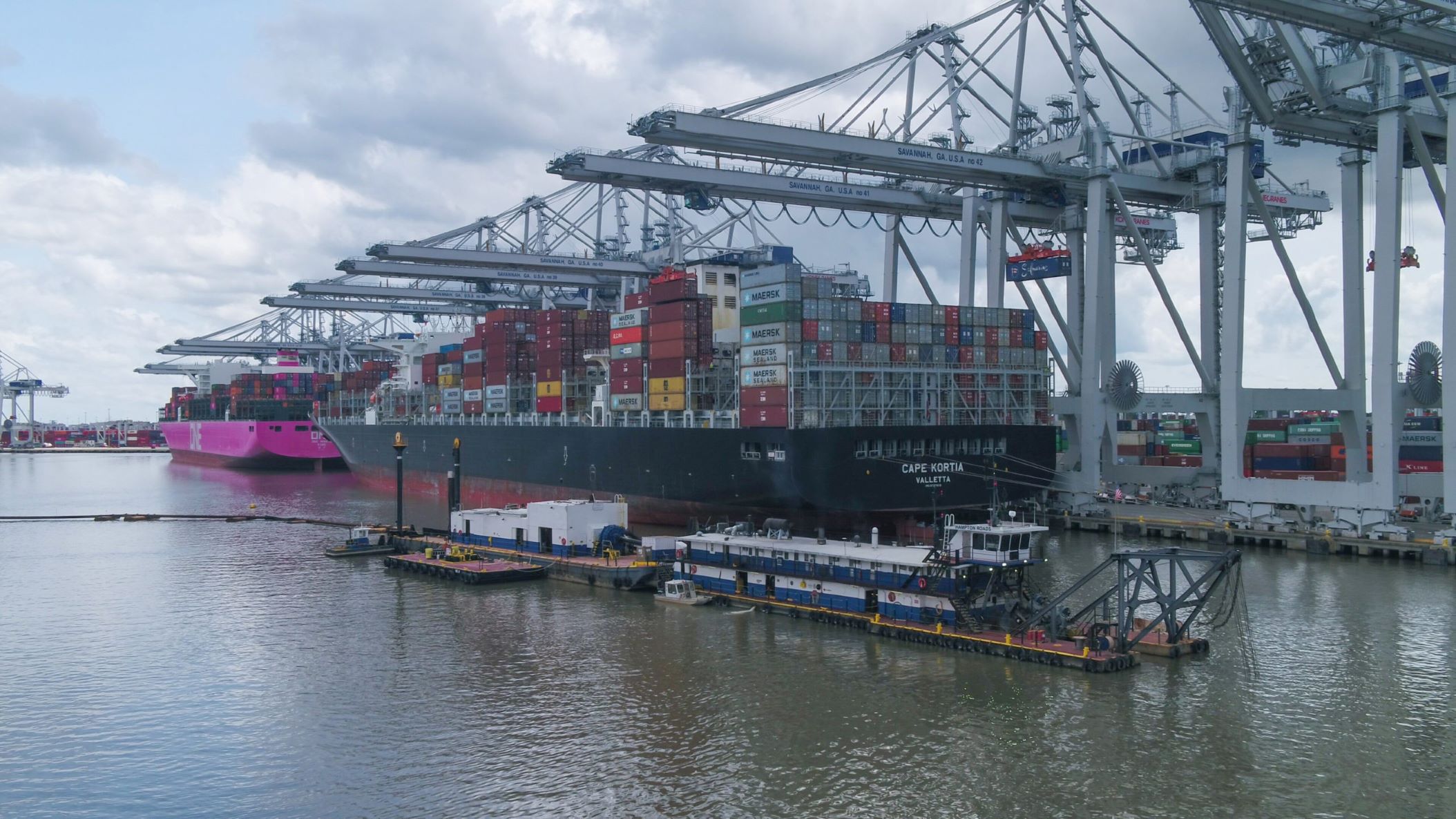 Dredging vessels alongside a massive container vessel docked at a port with large cranes overhead.