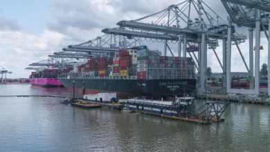 Dredging vessels alongside a massive container vessel docked at a port with large cranes overhead.