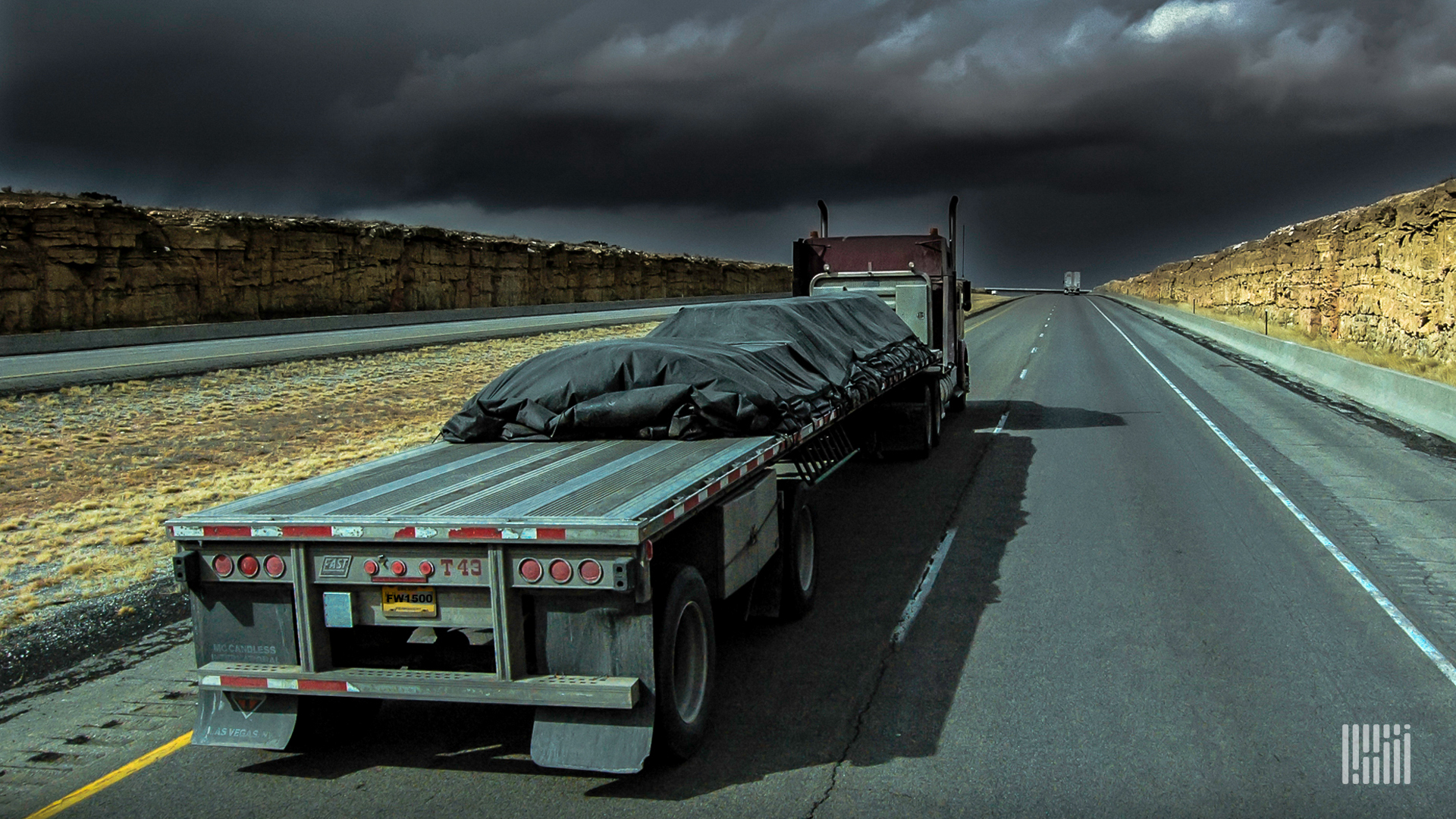 Flatbed tractor-trailer heading down highway with dark thunderstorm cloud across the sky.