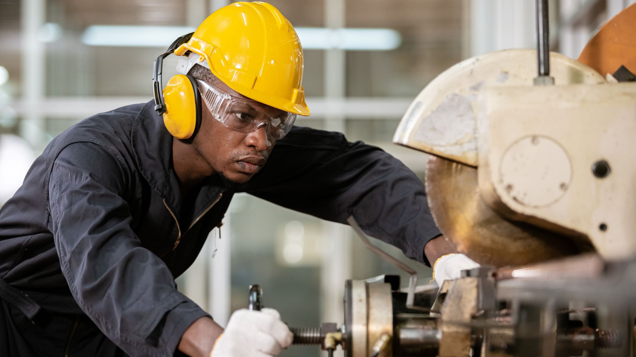 A black male wears soundproof headphones and yellow helmet working in an iron-cutting factory.