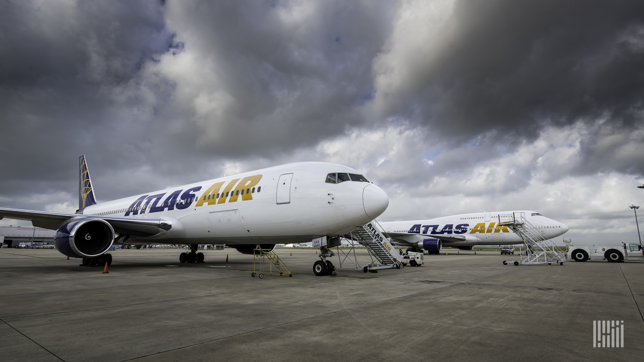 Two white passenger jets with blue accents sit on tarmac under stormy clouds.