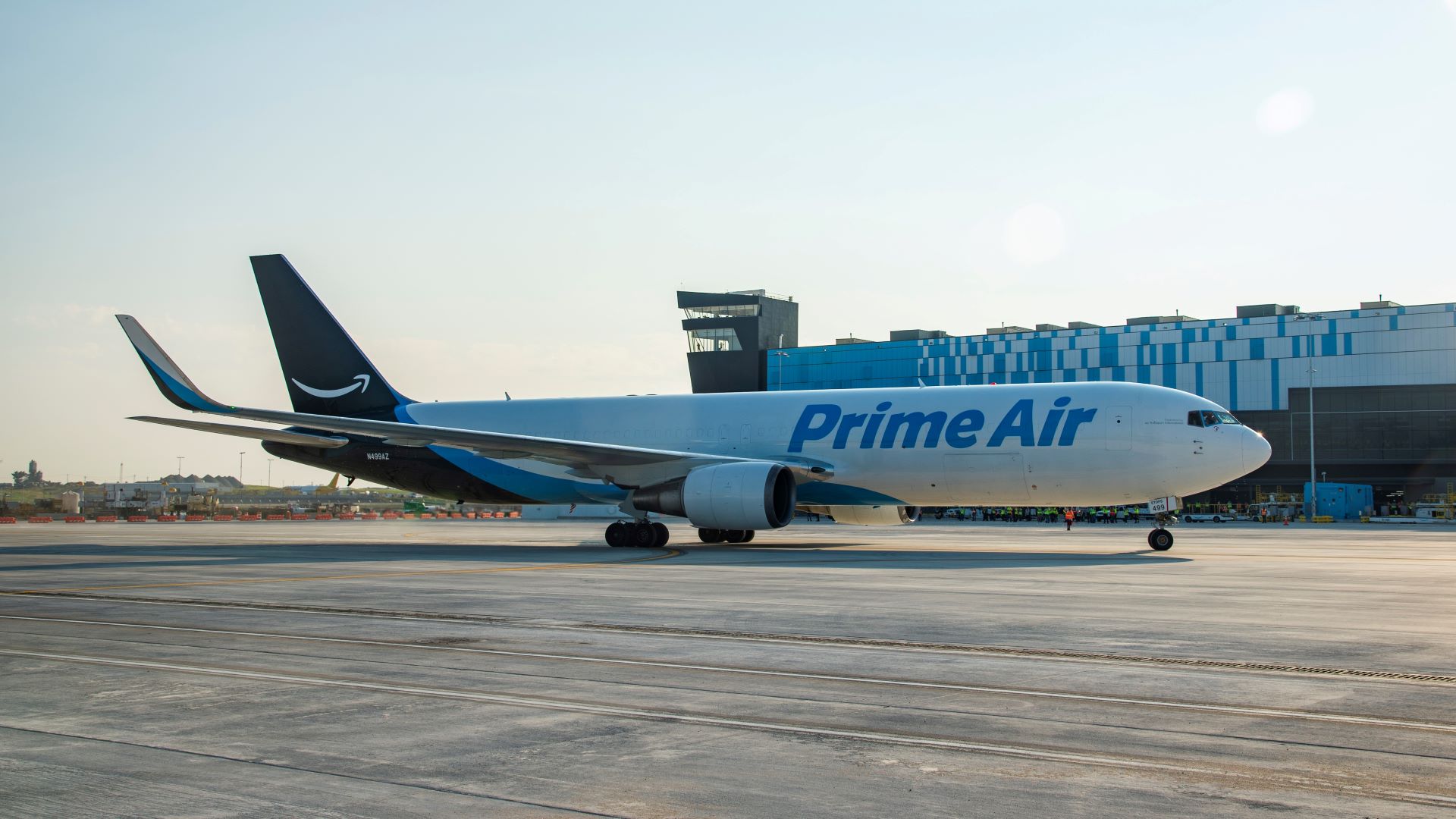 A white Amazon Prime jet with light blue accents rolls in front of a cargo building.
