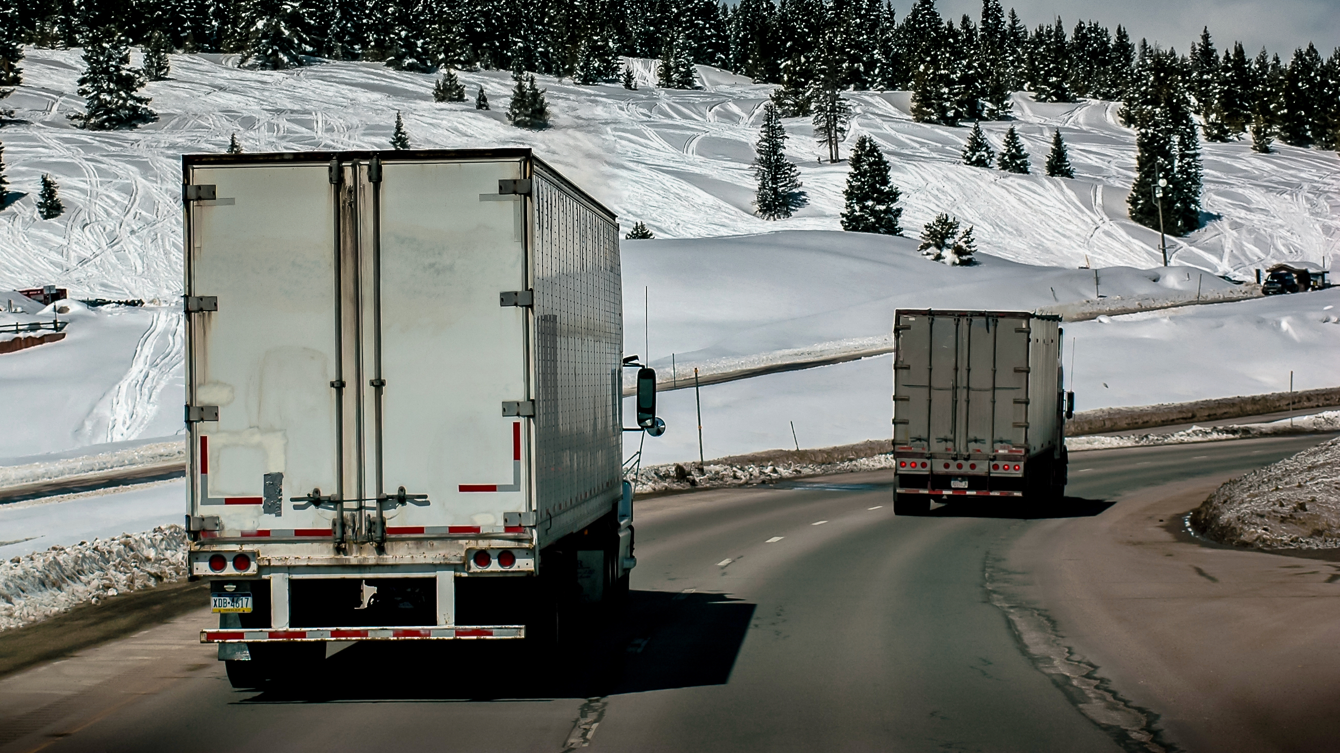 Trucks on the highway seen from behind in the Denver area with snow and trees in the background.