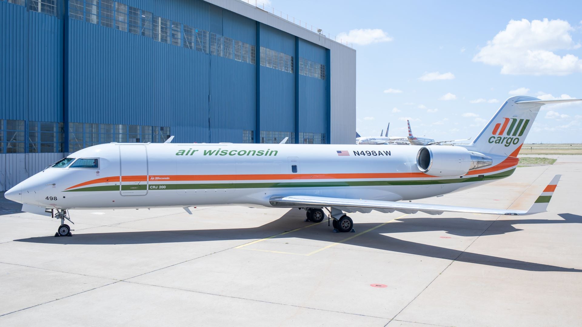 A small Air Wisconsin cargo jet sits on tarmac next to big hangar on bright day.