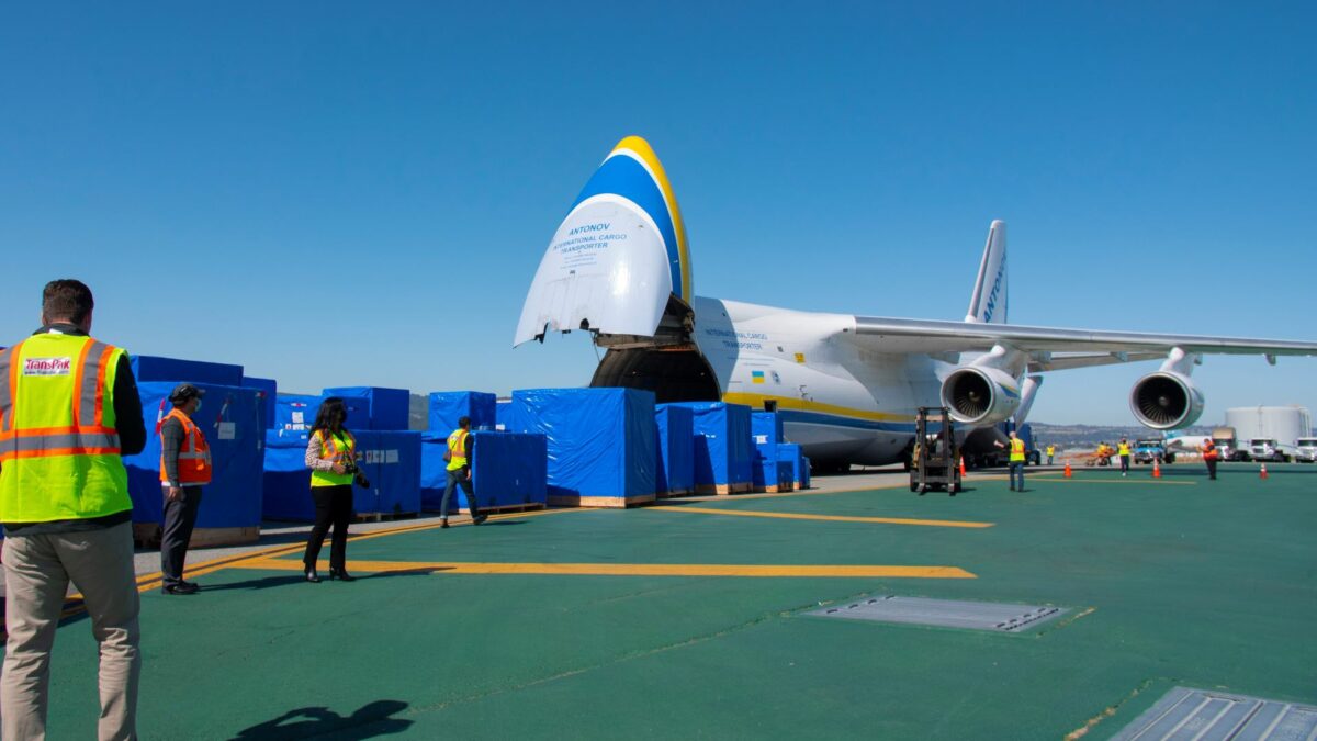 Large crates covered with blue tarps lined up in front of a nose-loading air cargo jet on a clear blue day.