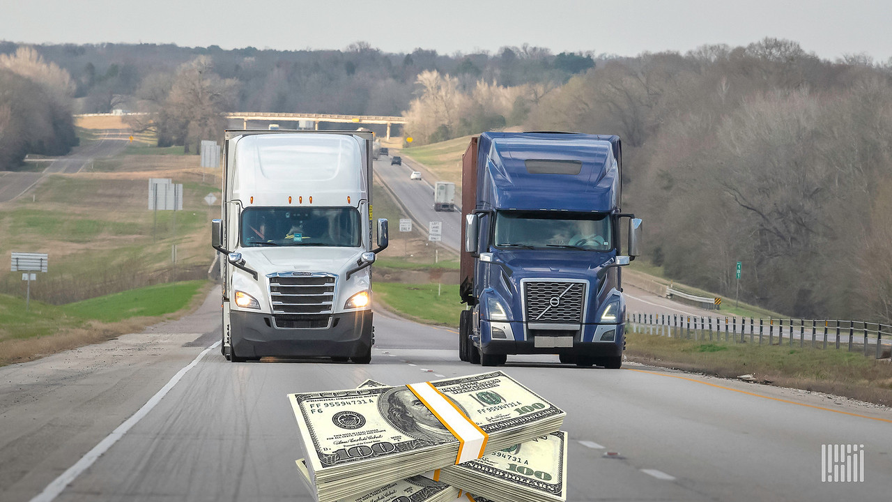 Two tractor-trailers, one white and one blue, travel next to each other on the road, where an image of a bundle of $100 bills is seen.
