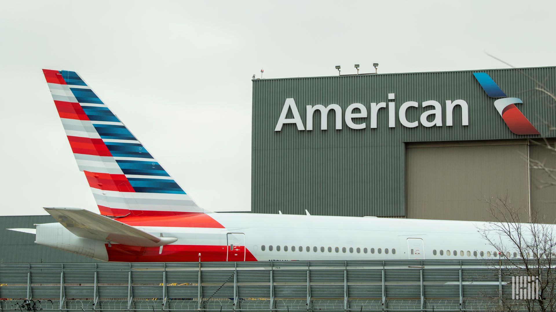 Tail end of a red, white and blue American Airlines jet close to an American hangar with the brand name on front.