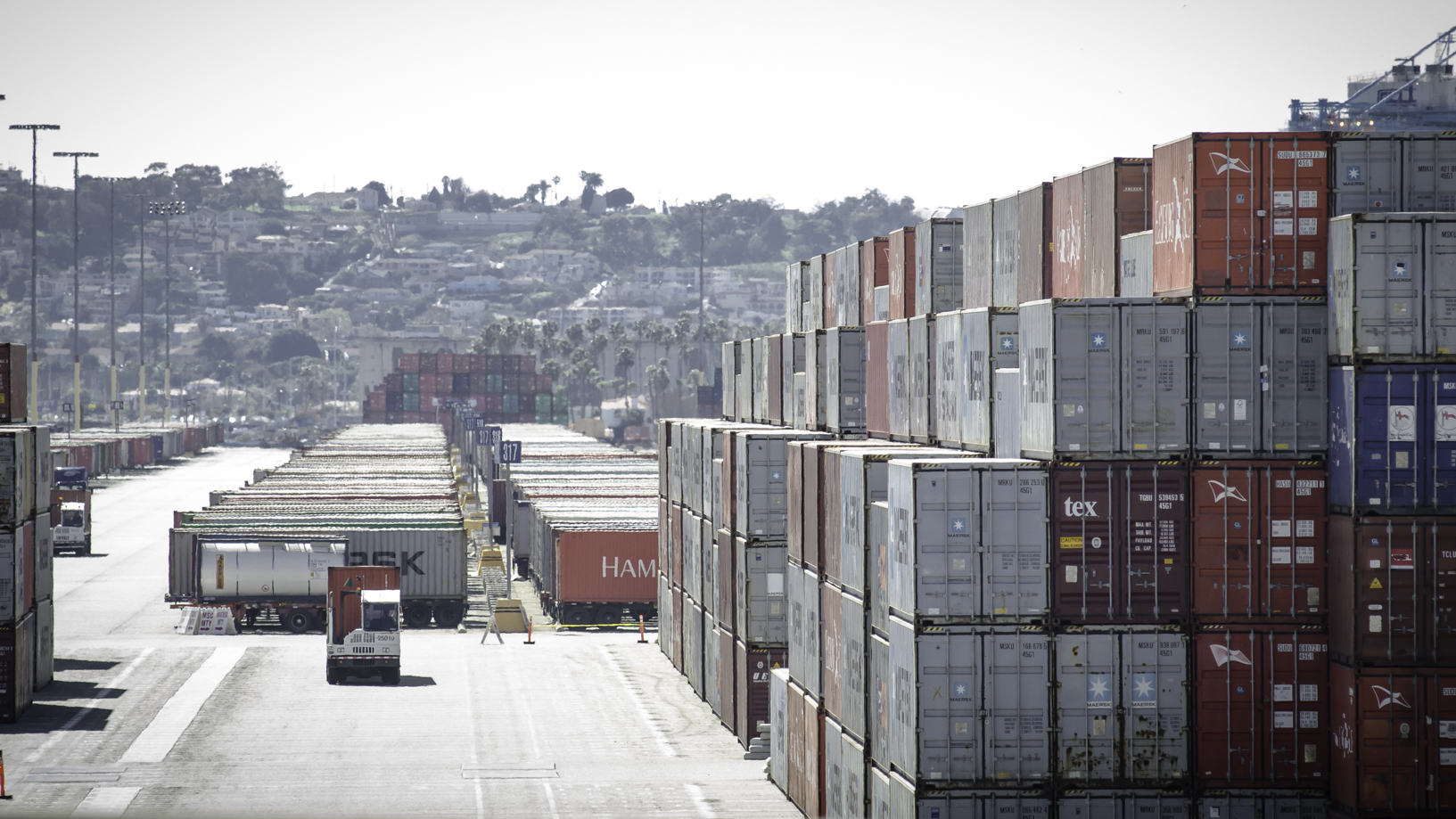 Stacks of containers at a port, with trucks in the background.