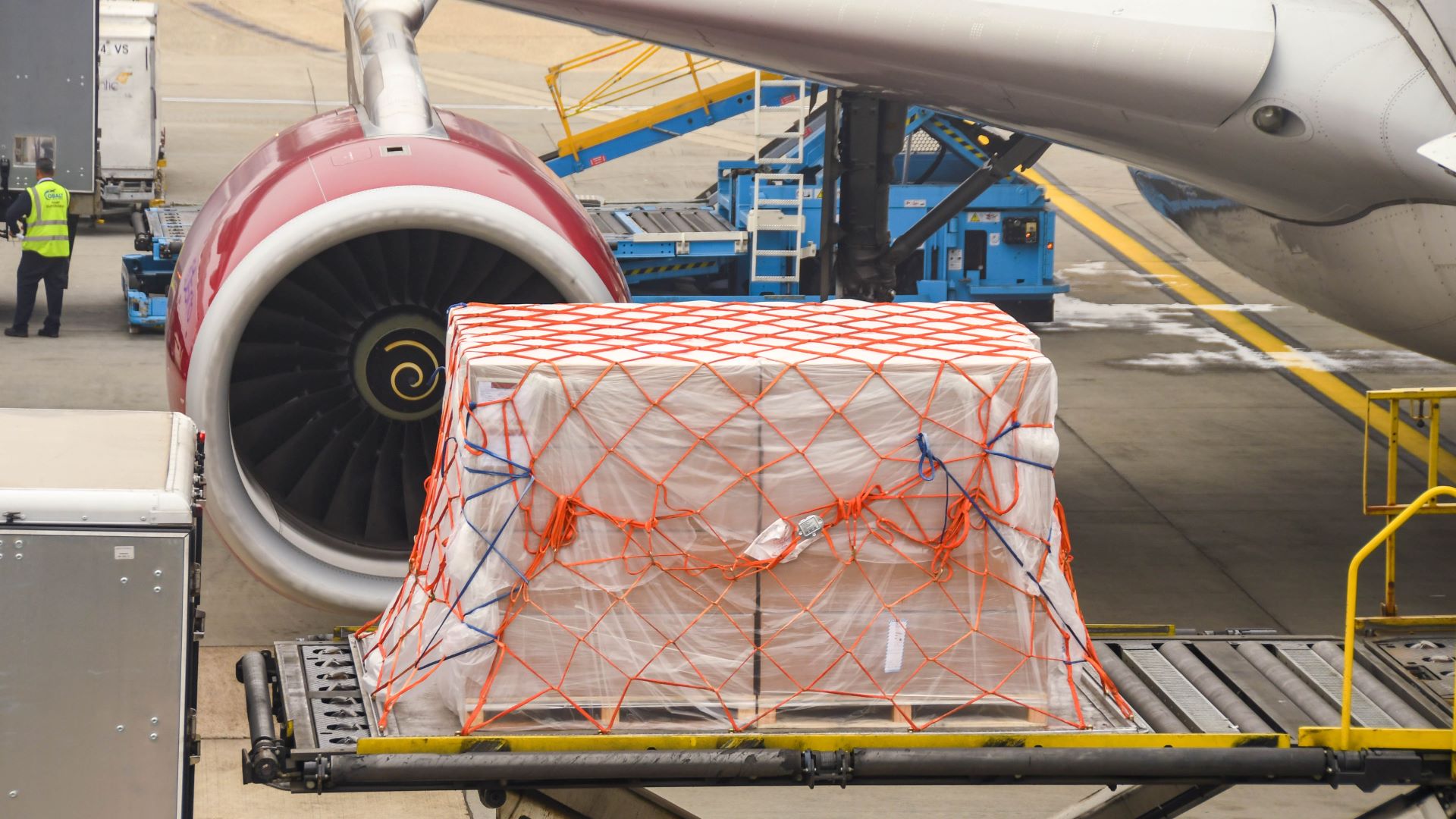 A close up of a cargo pallet wrapped in plastic on a lift ready to go in the old of an aircraft, waiting in front of jet engine.