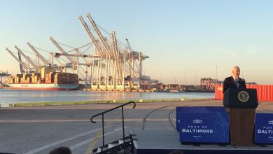 President Biden speaking at podium with port cranes and ship in the background.