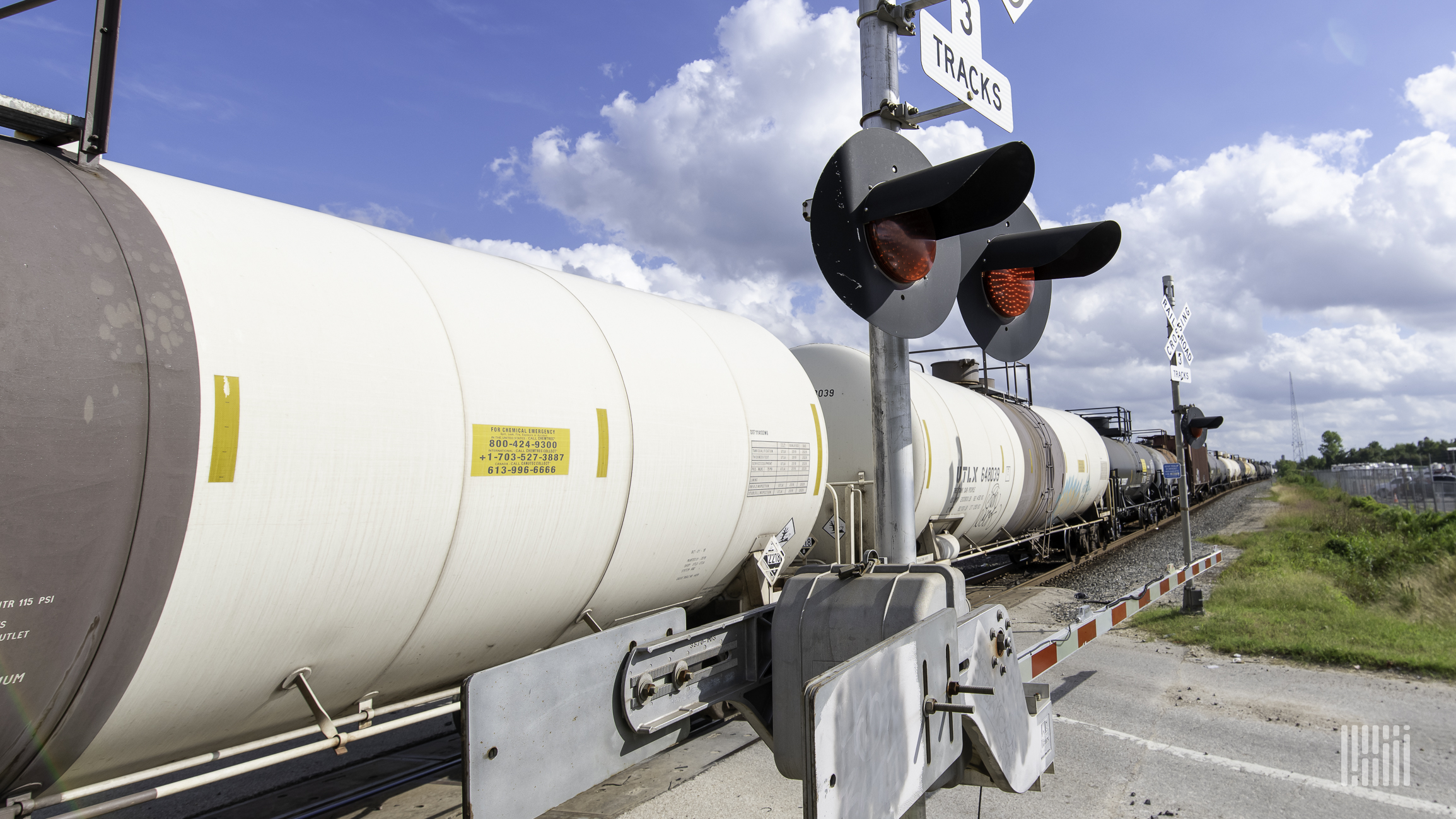 A photograph of a train of tank cars rolling through a rail crossing.