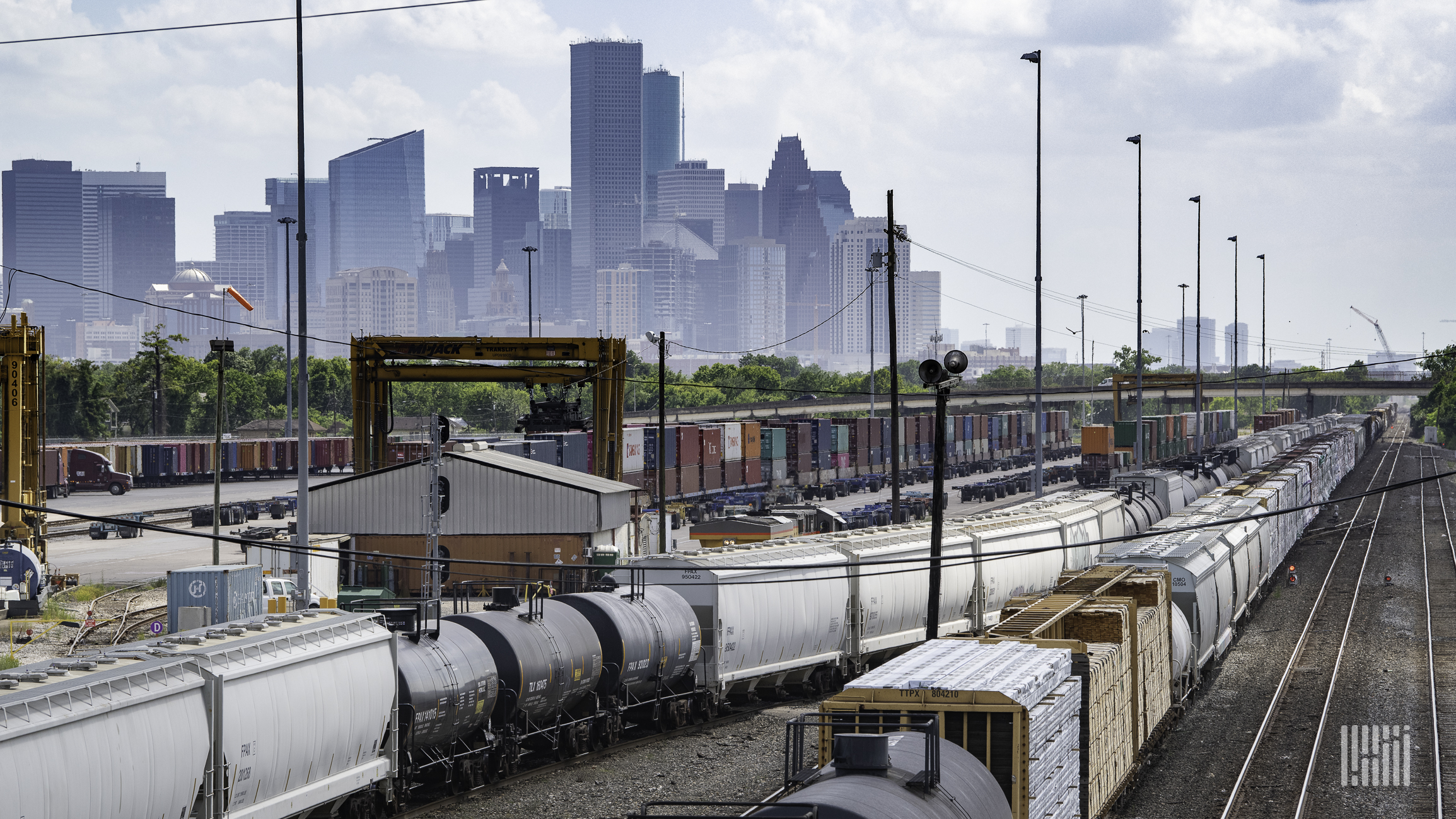 A photograph of railcars parked in a rail yard. There is a city skyline in the distance.