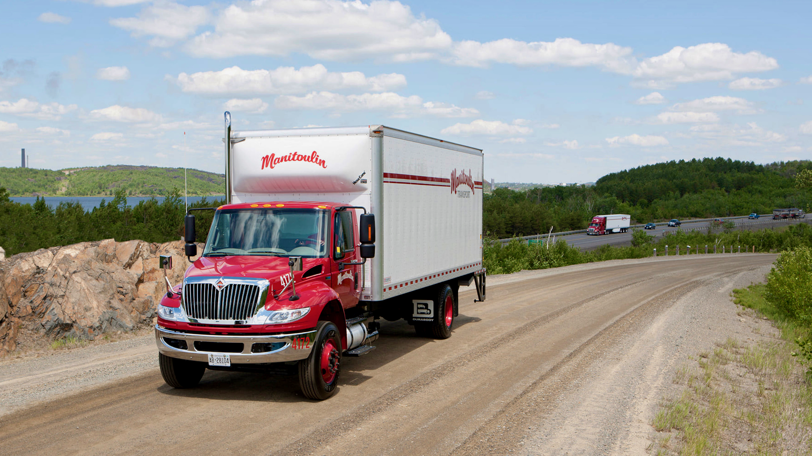 A red and white box truck of Manitoulin Transport on a dirt road with Manitoulin tractor-trailer on a highway the background