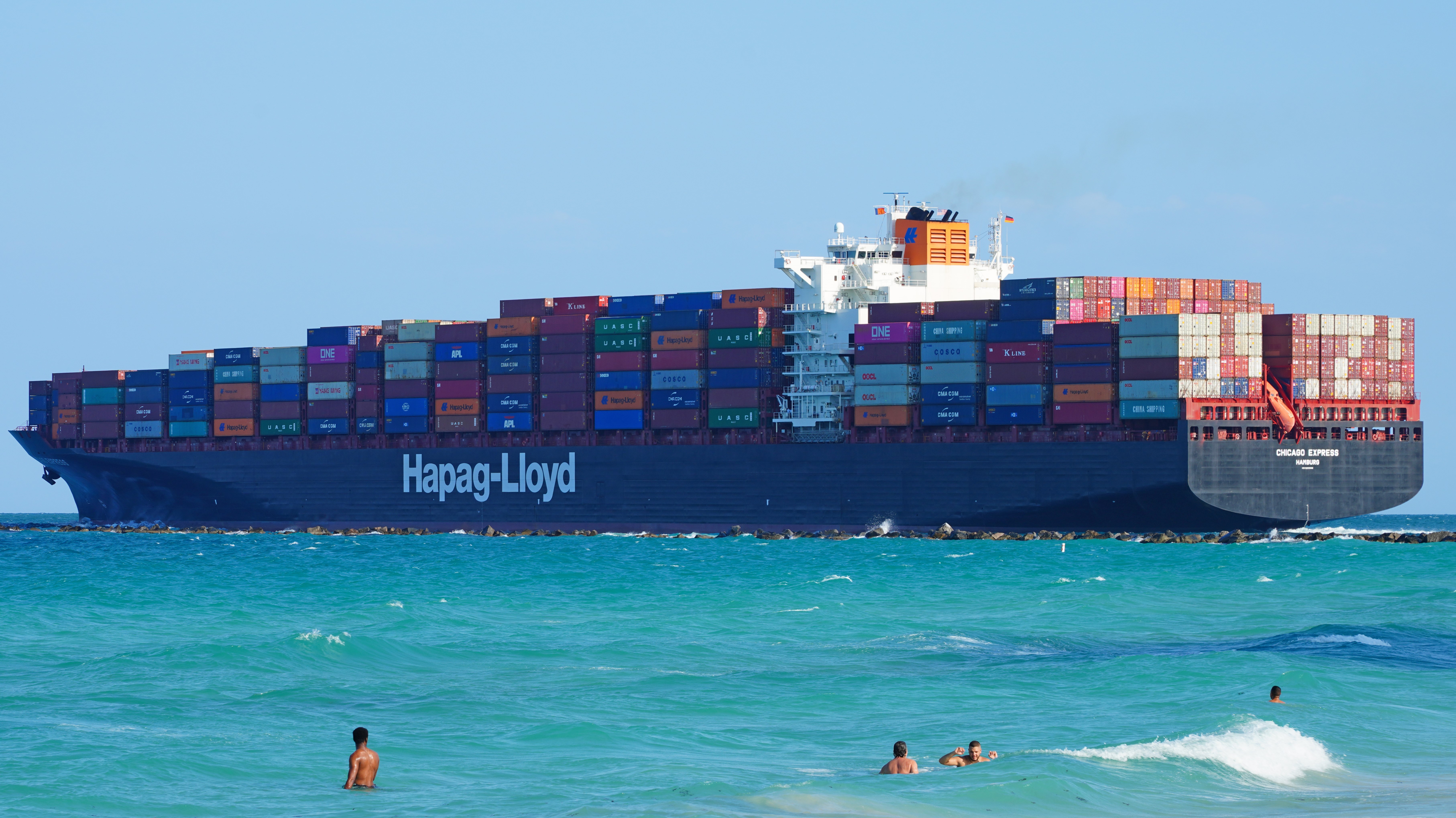 A large container vessel heads out to sea near beach on sunny day.