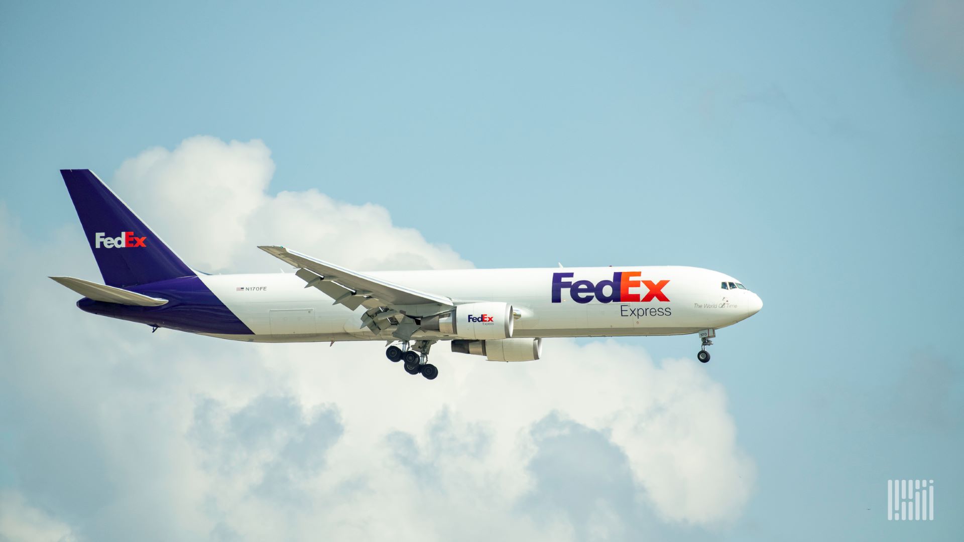 A white FedEx cargo jet with a purple tail flying across blue sky on landing approach.