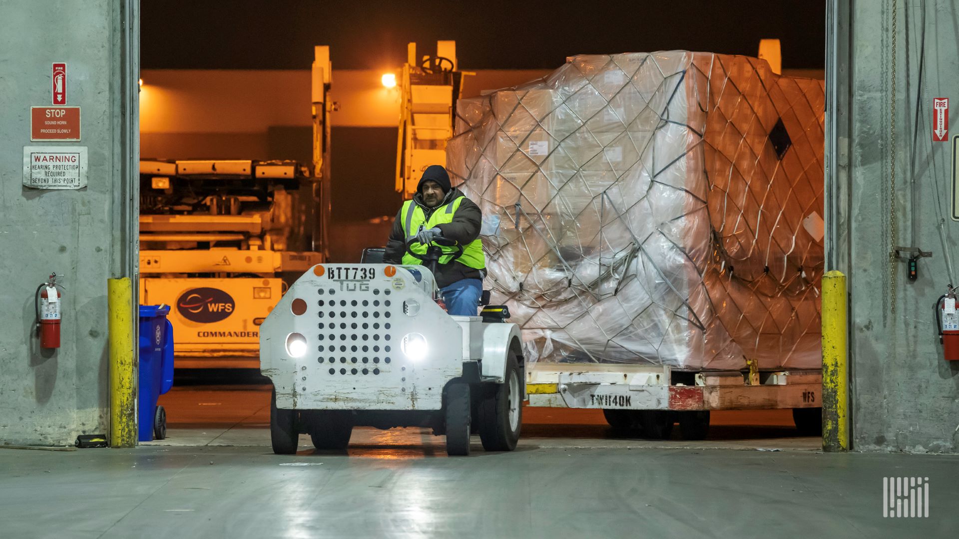 A motorized tug pulls a large air cargo pallet into a warehouse.