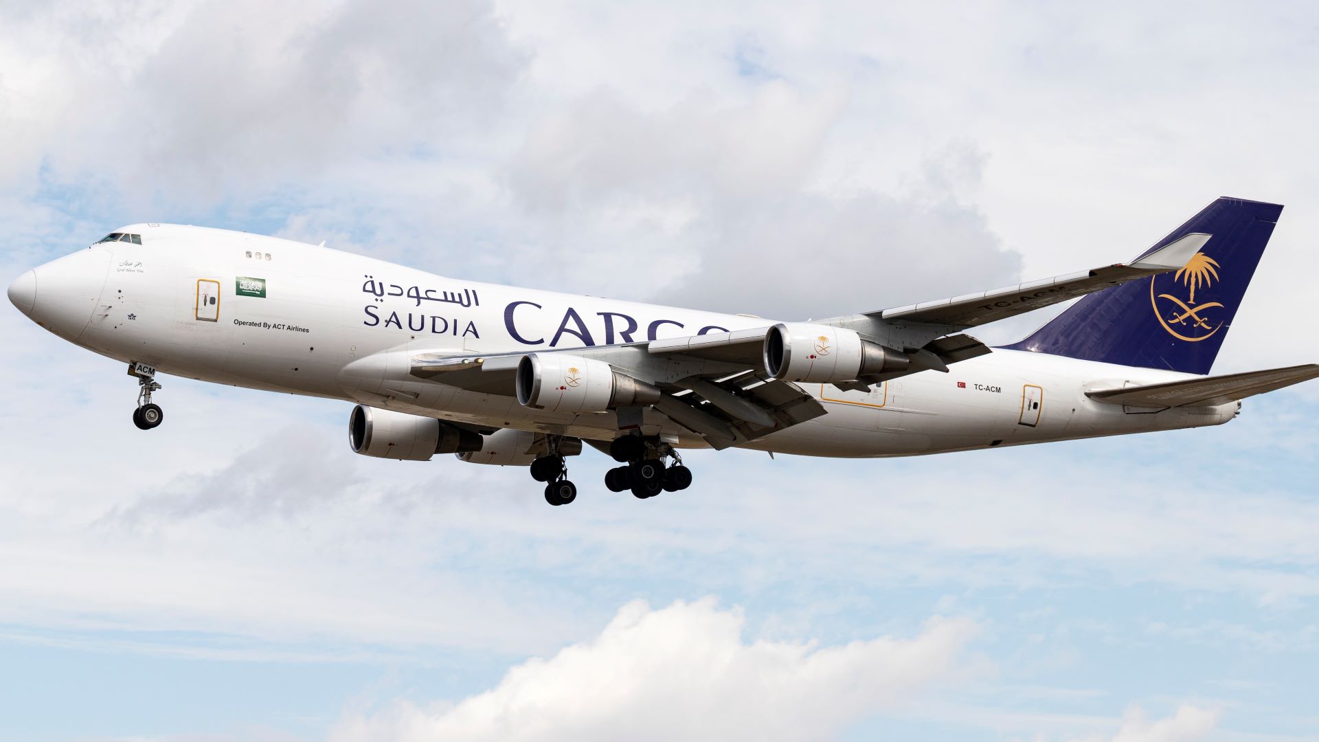 A white jumbo jet with teal green tail and Saudia Cargo on side comes in for landing with wheels down. View is looking up at plane in sky.