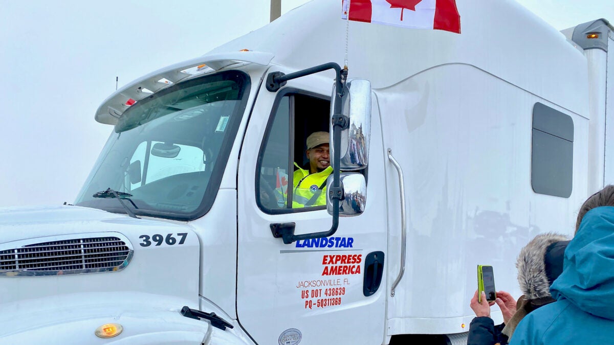 A truck driver in a white truck with a Canadian flag flying poses for a picture. 