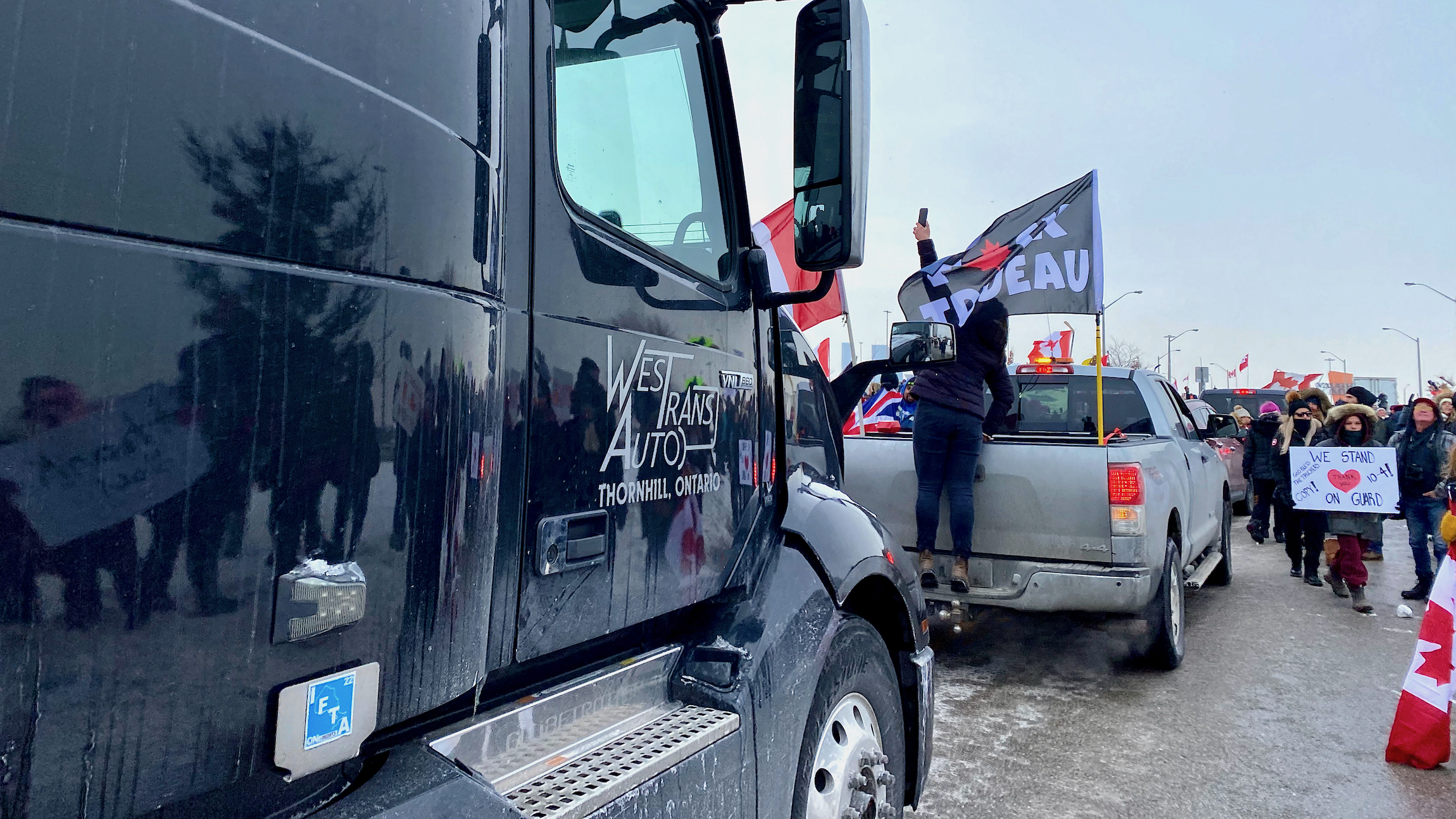 A semi truck moves forward as people holding signs and Canadian flags cheer on.