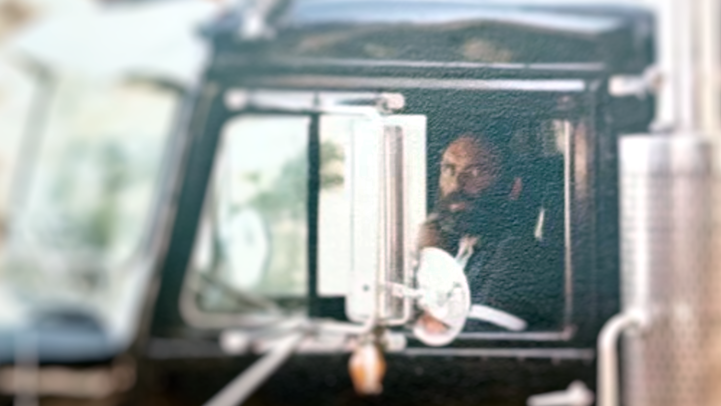 An old image of a truck driver, John Kelly, with a beard sitting in a truck